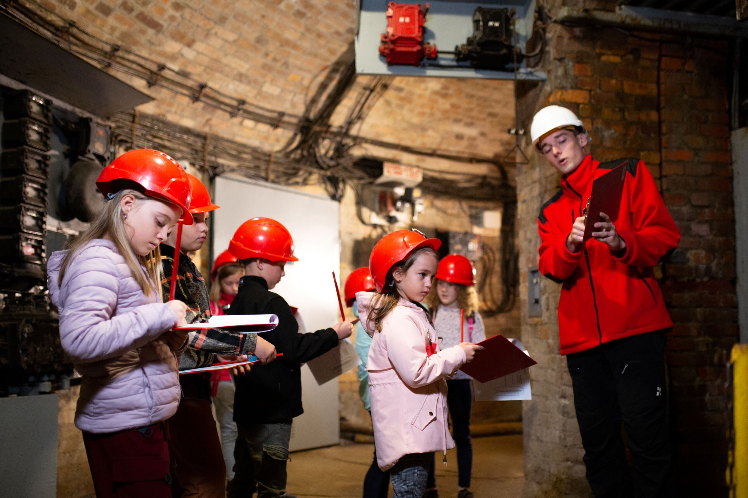 Grupa dzieci w czerwonych kaskach i z podkładkami do notowania patrzy na przewodnika w białym kasku.