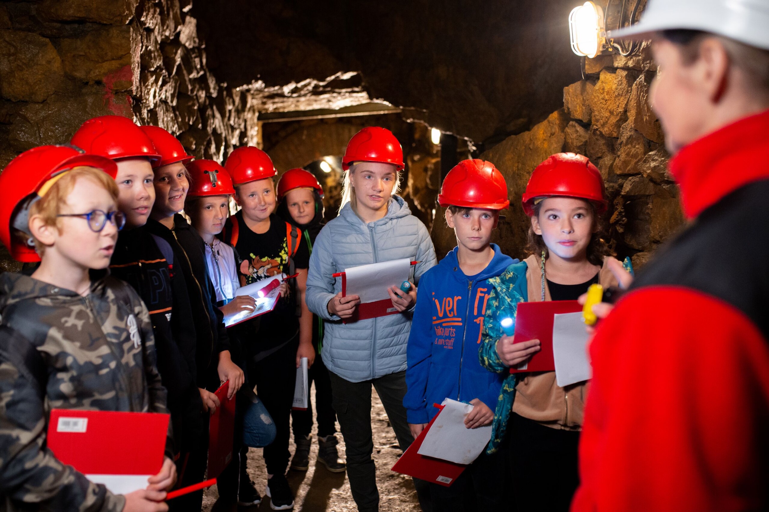 Grupa dzieci w czerwonych kaskach i z podkładkami do notowania patrzy na przewodniczkę w białym kasku.