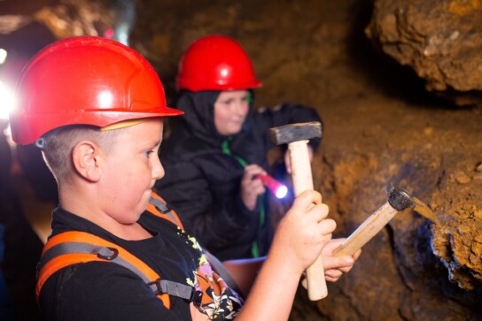 Chłopiec w czerwonym kasku stukający kilofkami w skały podczas lekcji edukacyjnej w podziemiach Zabytkowej Kopalni Srebra