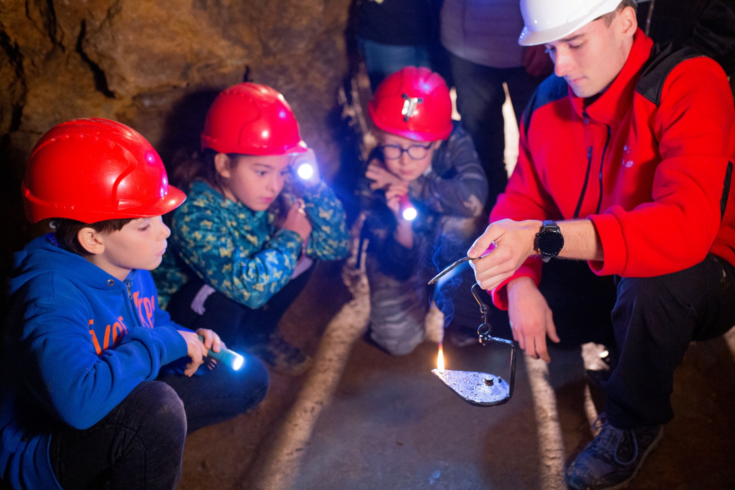 Przewodnik trzyma zapalony kaganek, obok niego trójka dzieci przygląda się płomieniowi